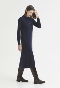 Femme portant une robe midi bleu marine à manches longues avec des boutons, marchant et touchant ses cheveux, associée à des bottes noires montantes.