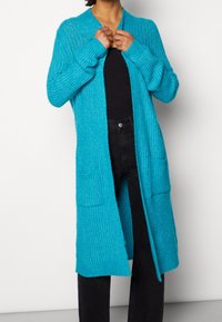 Lungo cardigan turchese con motivo a maglia testurizzata, apertura frontale, due tasche anteriori e polsini a costine. Indossato sopra un top nero.