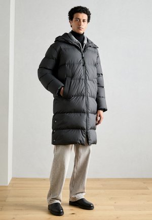 Dunkelgraue Daunenjacke mit Kapuze, Reißverschluss und seitlichen Taschen; kombiniert mit hellbeigen Hosen und schwarzen Schuhen auf einem Holzboden.
