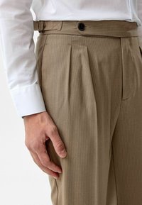 Beige geplooide broeken met een krijtstreep patroon, voorzien van een knoopsluiting en riemlussen, gedragen met een wit overhemd met lange mouwen.