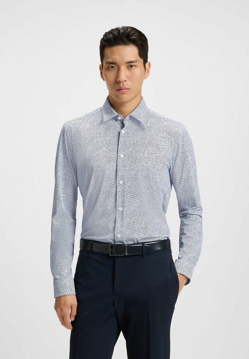 Camicia button-up in azzurro chiaro con un sottile motivo a pois. Maniche lunghe con polsini a bottone, abbinata a pantaloni scuri e una cintura.