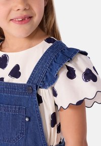 Salopette en denim avec des bretelles à volants, associée à un haut blanc avec un motif de coeurs navy. La salopette comprend une poche avant.