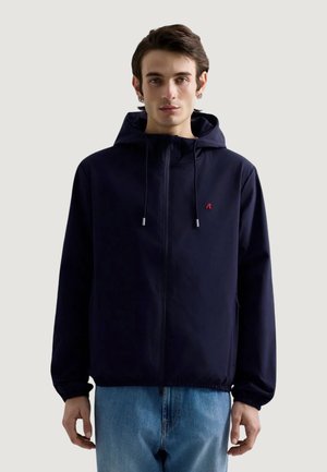 Ung mand iført en navy hættetrøje med lynlås og lyseblå jeans, stående mod en ensfarvet lys baggrund.