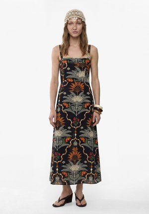 Frau trägt ein ärmelloses schwarzes Maxikleid mit orange-grünen Blumenmustern, einen beigen Häkelhut, braune Keilsandalen und Armreifen.