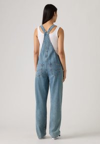 Τζιν ολόσωμη φόρμα σε μεσαίο μπλε χρώμα με ρυθμιζόμενους ιμάντες, λεπτομέρειες σε πίσω τσέπη και άνετη εφαρμογή. Φοριέται πάνω από λευκό αμάνικο τοπ.