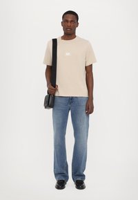 Beige bomulds t-shirt med trykt logo, parret med lyseblå denimjeans. Modellen bærer en lille sort crossbody-taske og har sorte sko på.