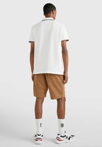 Polo bianco con colletto a righe rosse e blu, abbinato a pantaloni corti marroni. Calzature bianche e blu con accenti neri e logo. Calzini bianchi.