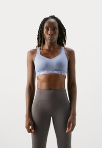 Mujer atlética con el cabello largo trenzado, vistiendo un sujetador deportivo Nike de color azul claro y leggings gris de cintura alta, de pie frente a un fondo blanco liso.