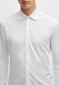 Chemise à manches longues blanche en tissu lisse, avec un col boutonné et un logo discret sur la poitrine, dotée de fermetures à boutons claires.