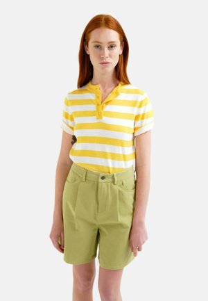 Mujer con cabello rojo que lleva una camiseta de manga corta a rayas amarillas y blancas y pantalones cortos de cintura alta de color verde claro, de pie frente a un fondo liso.