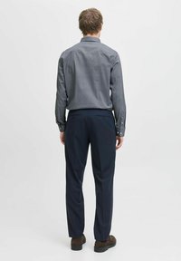 Bărbat în picioare, întors cu spatele, purtând o cămașă cu model, pantaloni închisați la culoare și pantofi maro, pe un fundal alb simplu.