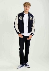 Chaqueta varsity azul marino y crema con parches bordados, puños acanalados y cierre de cremallera, combinada con vaqueros negros y zapatillas azules.
