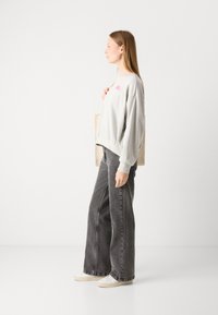 Grijze sweater met roze borduursel, gecombineerd met brede grijze denim jeans. Model houdt een beige schoudertas vast. Platte, casual schoenen worden gedragen.