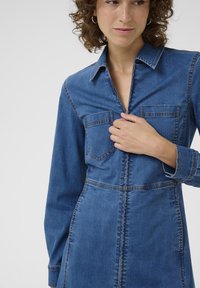 Rochie din denim de culoare albastru mediu, cu talie ajustată, mâneci lungi, guler și două buzunare frontale. Prezintă cusături contrastante și fermoar în față.