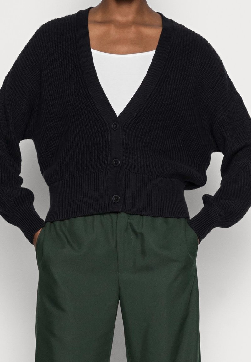 Cardigan noir à côtes avec un décolleté en V profond, une fermeture à boutons à l'avant et des épaules tombantes, associé à un pantalon vert ample.
