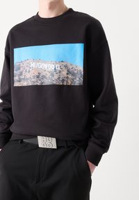 Černá mikina s modrým obdélníkovým motivem s nápisem "HUGOWOOD" na kopci. Nosit se černými kalhotami a stříbrným páskem s nápisem "HUGO".