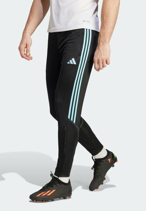 Mann trägt schwarze Adidas-Trainingshose mit hellblauen Streifen und schwarze Stollenschuhe mit orangenen Details vor schlichtem Hintergrund.
