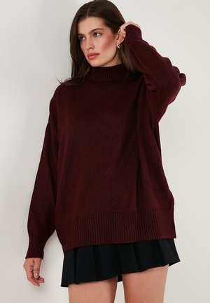Pulover oversized bordo cu guler înalt, cu margini și manșete cu serpentine, realizat dintr-un material moale și texturat. Stilizat cu o fustă plisată neagră.