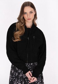 Giacca cropped in suede nero con zip frontale, polsini a coste e tasca sul petto. Indossata sopra una gonna a fantasia floreale.
