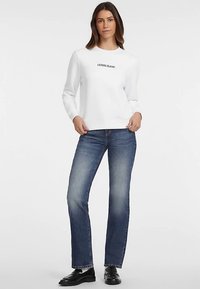 Valkoinen collegepaita, jossa "GUESS JEANS" mustalla painettuna, yhdistetty tummansinisiin farkkuihin. Mallilla on mustat kengät, ja hän seisoo vaaleaa taustaa vasten.