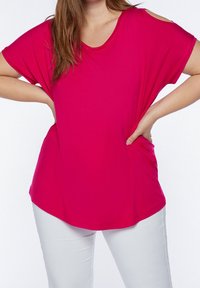 Woman wearing a bright pink cold-shoulder top and white pants, hands resting on hips, against a plain white background.