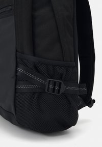 Mochila negra con un bolsillo lateral de malla texturizada asegurado con un broche negro y acentos en gris a rayas, con un diseño de cuerpo de tela suave.