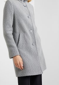 Grått texturerat ullkappa med stora knappar, krage och sidofickor, med rak skärning och långa ärmar.