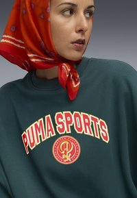 Zelená mikina s logom "PUMA SPORTS" v tučnej červenej a žltej farbe. Má okrúhly výstrih a textúrovanú tkaninu. Okolo krku je viazaný oranžový šatka so vzorom.