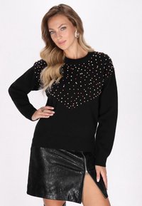 Pulover negru cu mărgele colorate pe față, asortat cu o fustă lucioasă din piele sintetică neagră, având un detaliu cu fermoar pe lateral.