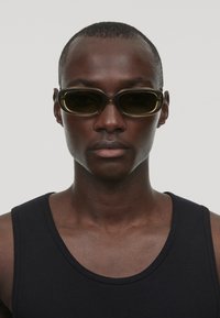 Des lunettes de soleil noires avec une forme ovale élégante et des verres teintés, encadrées d'une subtile couleur olive. Portées par une personne en débardeur noir.