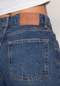 Acercamiento a la parte trasera de unos jeans de mezclilla azul con un parche de cuero etiquetado "BDG" en la cintura, llevados con una camiseta cropped blanca.