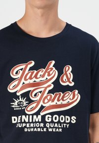 Tamnoplava pamučna majica s bijelim i crvenim grafičkim printom koji sadrži "Jack & Jones", grafiku suncu te natpis "Denim Goods, Superior Quality."