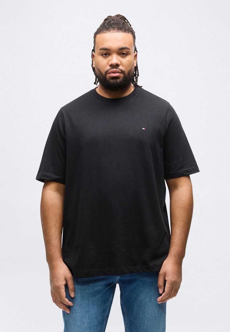 Άνδρας με πλεξουδάκια στα μαλλιά και γενειάδα, φορώντας μαύρο μπλουζάκι και μπλε τζιν, στέκεται μπροστά σε απλό ανοιχτό γκρι φόντο.