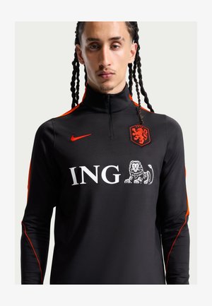 Homme aux cheveux tressés portant une tenue de sport noire et orange avec le logo ING, blason de lion, short noir, chaussettes noires et baskets noires, debout.