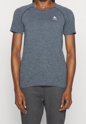 Osoba ubrana w dopasowaną, krótkim rękawem, ciemnoszarą teksturowaną sportową koszulkę i dopasowane ciemnoszare spodnie, stojąca na jednolitym tle.