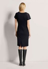 Schwarzes Kleid mit kurzen Spitzenärmeln, taillierte Passform, knielang, mit Schlitz hinten, getragen mit knielangen schwarzen Stiefeln, glatter Stoff.