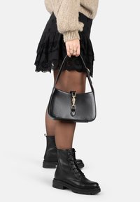 Vrouw in een zwarte kanten rok, doorschijnende panty's, zwarte veterschoenen, een beige gebreide trui, en met een zwarte leren handtas met gouden hardware.