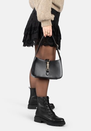 Femme portant une jupe en dentelle noire, des collants transparents, des bottes en cuir noir à lacets, un pull en tricot beige, et tenant un sac à main en cuir noir avec des accessoires en or.