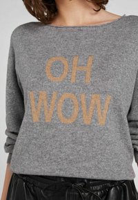 Maglione grigio a maglia con maniche arrotolate e testo beige "OH WOW" indossato con pantaloni neri con cordino.