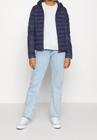 Chaqueta de plumas navy con capucha, puños de canalé y bolsillos con cremallera, combinada con una sudadera gris y jeans azul claro, y zapatillas blancas.