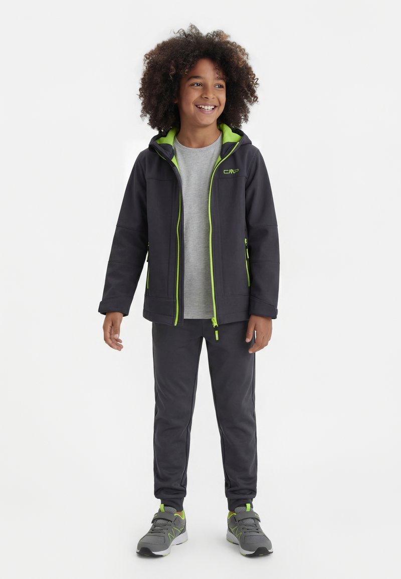 Usmievajúce sa dieťa s kučeravými vlasmi, oblečené v tmavej kapucňovej bunde so svetlozeleným lemovaním, šedej tričke, tmavých nohaviciach a sivých teniskách, stojí čelom vpredu.