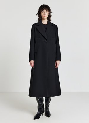 Manteau long en laine noire avec un col cranté, fermeture à boutons sur le devant et deux poches latérales. Porté avec des bottes noires brillantes arrivant au genou.