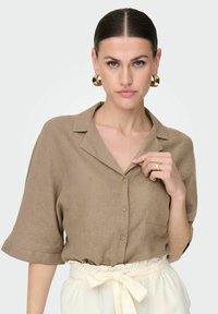 Camicia button-up in lino marrone con colletto, maniche corte e una tasca frontale, abbinata a pantaloni chiari con cintura annodata.