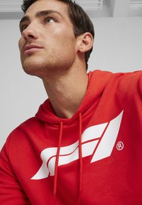 Rød bomulds-hoodie med et stort hvidt logo foran og snoretræk ved halsudskæringen. Glat tekstur med en afslappet pasform.