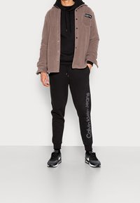 Hombre vistiendo una chaqueta de forro polar color topo con botones sobre una sudadera negra y pantalones de chándal negros de Calvin Klein Jeans, con zapatillas negras de Nike.