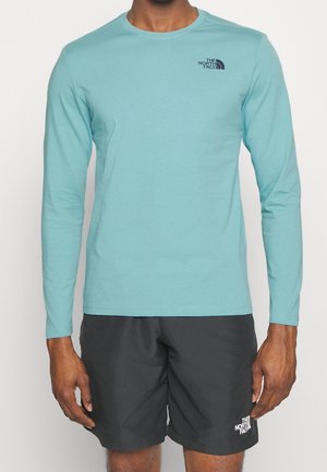 Mann trägt hellblaues Langarmhemd und schwarze Shorts, beide mit dem The North Face-Logo, steht vor einem einfarbigen hellen Hintergrund.