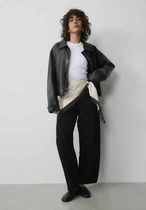 Schwarze, kurz geschnittene Lederjacke, weißes T-Shirt, weite schwarze Hose und ein cremfarbener Seidenschal mit Spitzenbesatz. Schwarze Schuhe vervollständigen das Outfit.