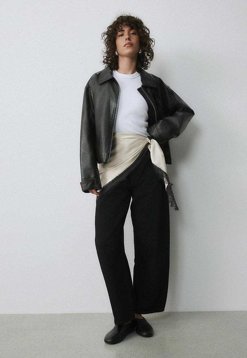 Černá kožená krácená bunda, bílé tričko, široké černé kalhoty a krémový hedvábný zavinovací šátek s krajkovým lemem. Černé boty doplňují outfit.