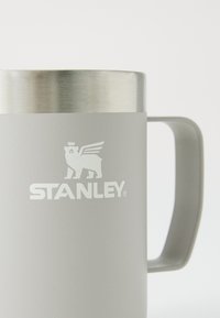 Caneca isolada cinza com um aro em aço inoxidável, apresentando um proeminente logótipo branco "STANLEY" e uma pega; textura mate suave.