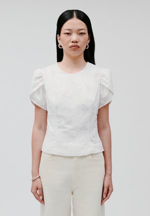 Blusa bianca dalla texture morbida con maniche a sbuffo, collo rotondo e vita aderente, abbinata a pantaloni beige chiaro. Dettagli di un sottile motivo floreale visibili.